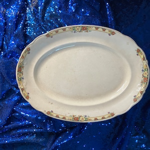May include: A white oval serving platter with a floral border in shades of yellow, orange, and red. The platter is on a blue sequined background.