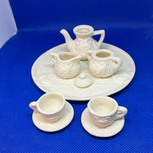 May include: A miniature white porcelain tea set, comprising a teapot, creamer, sugar bowl with lid, and two teacups with saucers. The set is arranged on a round, embossed serving plate. Ideal for dollhouses or miniature collections.