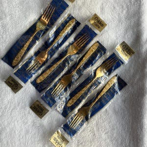 以下が含まれることがあります： 個別に包装された、いくつかの金色のフォーク。フォークは柄に装飾的な模様があり、青と透明のプラスチックで密封されています。各パッケージには「Golden Ware」と書かれた金色のラベルが付いています。