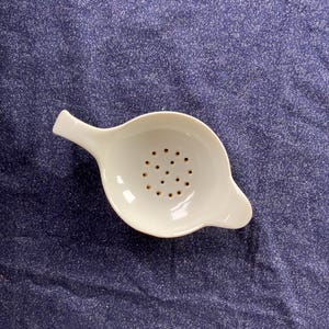 May include: A white ceramic tea strainer with a handle and small holes in the bowl. The strainer is designed to sit over a cup to catch tea leaves. The tea strainer is on a blue textured surface.
