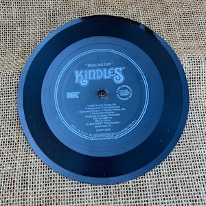 Könnte beinhalten: Eine schwarze Schallplatte mit dem Text "Music and Love" und dem Bandnamen "Kindles". Die Schallplatte hat ein graues Etikett mit zusätzlichem Text, darunter "IDEAL" und "EVATONE Soundsheets". Die Schallplatte liegt auf einer strukturierten, beigefarbenen Oberfläche.