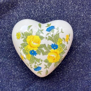 May include: A white heart-shaped ceramic trinket box featuring a floral pattern of yellow roses, small blue flowers, and green leaves. The surface is smooth and glossy, with the botanical design centered on the front of the heart-shaped object.