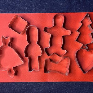 May include: A set of eight metal cookie cutters in various shapes, including a heart, a tree, a gingerbread person, a boot, and a bell. The cutters are on a red surface.
