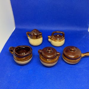 Puede incluir: Colección de miniaturas de cerámica para cocina, que incluye una tetera, una olla con asa y ollas con tapa. Las piezas son marrones y beige, con un acabado brillante. Sobre una superficie azul.