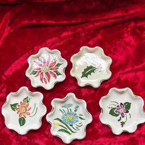 Könnte beinhalten: Fünf kleine, cremeweiße Keramikschalen mit gewelltem Rand und handgemalten Blumenmotiven. Jede Schale zeigt eine einzigartige botanische Illustration, darunter ein roter Weihnachtsstern, grüne Stechpalmenblätter sowie orange, blaue und violette Blüten.