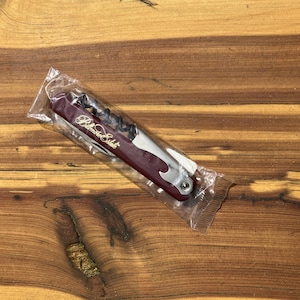 Peut inclure: Un ouvre-bouteille multifonctionnel bordeaux et argenté avec le texte "Belmore Estate" en lettres dorées, emballé dans du plastique transparent. L'outil comprend un tire-bouchon et un décapsuleur.