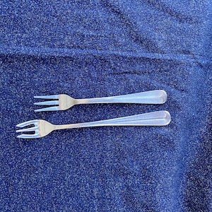 May include: Two silver forks with three tines each, resting on a textured, dark blue surface. The forks have a simple, elegant design, with a slightly curved handle. The image is a top-down view.