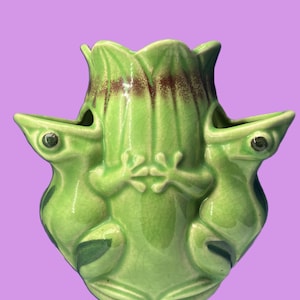 Puede incluir: Un jarrón de cerámica verde con tres figuras de rana. El jarrón tiene una parte superior en forma de tulipán con detalles marrones y aberturas en forma de rana. Las ranas tienen ojos oscuros y están sentadas.