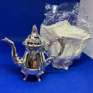 以下が含まれることがあります： 蓋とハンドルに詳細な花の模様が施された、小さく装飾的な銀色のティーポット。 ティーポットは湾曲した注ぎ口があり、3つの小さな足の上に立っています。 白い物体がティーポットの後ろのビニール袋に入っています。