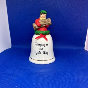 May include: A white ceramic bell with gold trim and the text "Bringing in the Yule Log." A figure in a red coat and green hat carries a log on top of the bell, which has a green wreath and red bow.