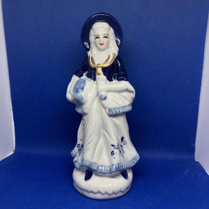 May include: A porcelain figurine of a woman in a long white dress with blue floral accents and a dark blue cloak. The figurine has a white bonnet and a gold-colored necklace. The base is white with a blue trim.