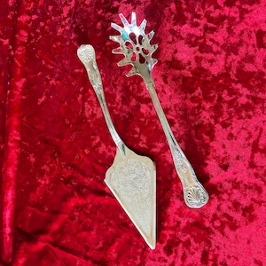 May include: Silver serving utensils on a red velvet surface. The set includes a pie server with a decorative handle and a pasta server with a slotted head. The utensils have ornate detailing.