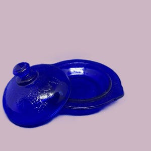 May include: A small, round, cobalt blue glass butter dish with a matching lid. The lid features a central knob handle and a textured surface pattern. The base is circular with a slightly raised rim, designed to hold the lid securely in place.