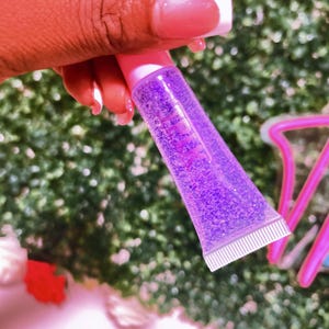 May include: A clear tube of purple lip gloss with glitter. The tube is pink and has a white cap.
