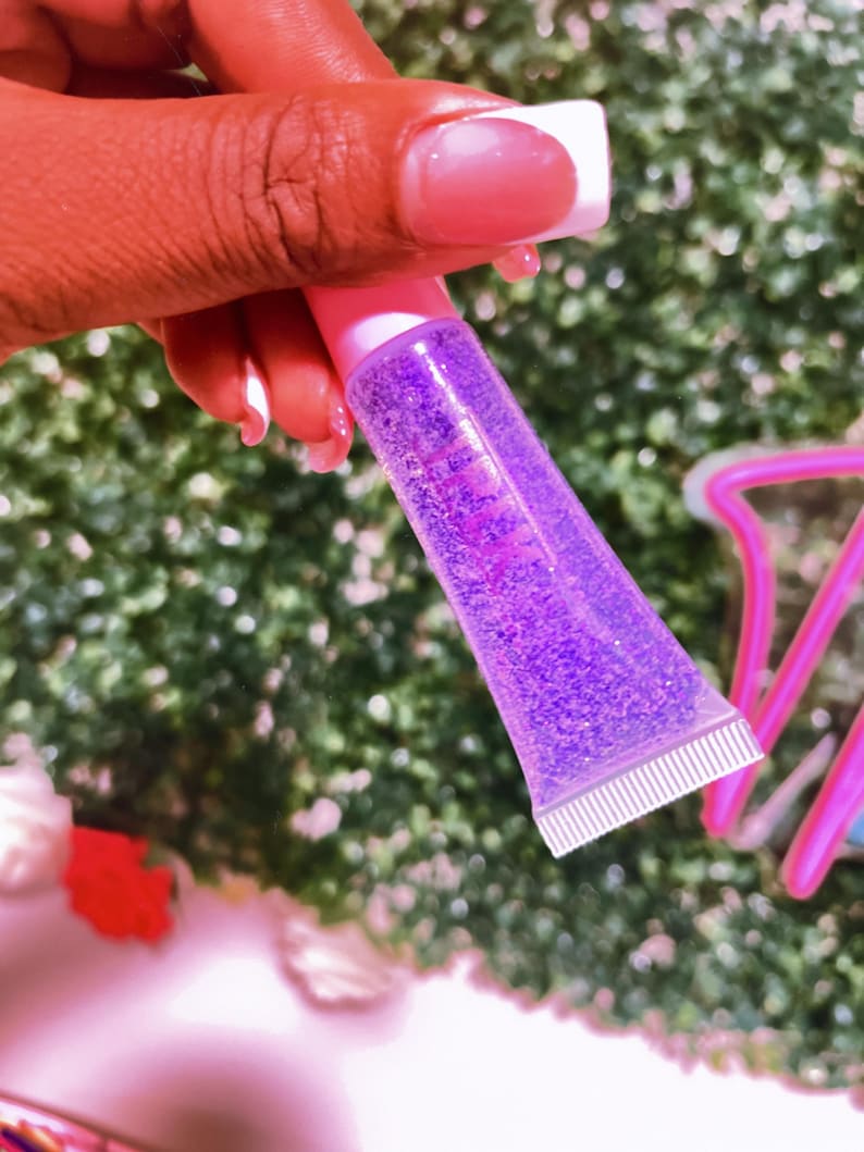 May include: A clear tube of purple lip gloss with glitter. The tube is pink and has a white cap.