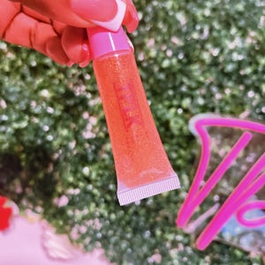 May include: A clear tube of pink lip gloss with a pink cap. The gloss is a light orange color with glitter.