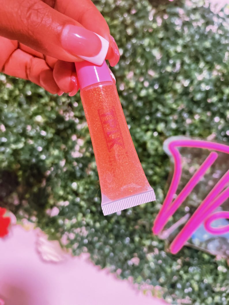 May include: A clear tube of pink lip gloss with a pink cap. The gloss is a light orange color with glitter.