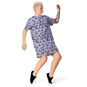 May include: A lavender t-shirt dress with a black and white abstract face pattern. The dress has short sleeves and a relaxed fit.