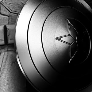 May include: Black and white image of a Captain America shield with a star emblem. The shield is metallic and has concentric circles. The image also shows a portion of a costume and utility belt. The text "AVENGERS ASSEMBLE" is visible.