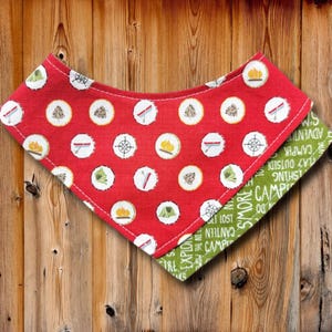 Op de afbeelding: Rode en groene bandana met een campingthema. De rode kant heeft witte cirkels met campingiconen zoals kampvuren, kompassen en tenten. De groene kant heeft de woorden "Camp" en "Explore" in het wit.