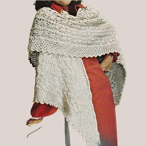 May include: A cream-colored crocheted shawl featuring an intricate openwork lace pattern with a scalloped edge. The shawl is draped over a solid rust-colored long-sleeved garment, showcasing the detailed texture and craftsmanship of the yarn work.
