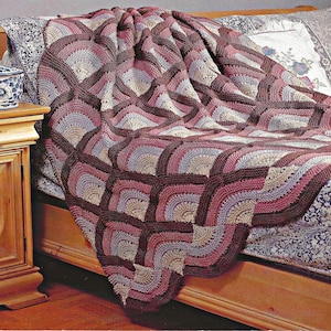 Könnte beinhalten: Eine handgefertigte, gehäkelte Decke mit einem geometrischen Muster. Die Decke zeigt ein sich wiederholendes Design aus Quadraten mit gebogenen Kanten in den Farbtönen Burgunder, Rosa, Lavendel und Beige. Die Decke ist über ein Bett drapiert und zeigt ihre Textur und ihr Design.