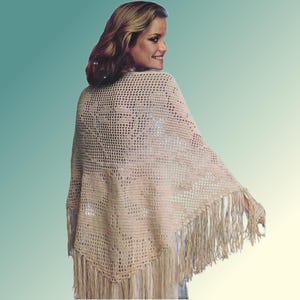 May include: A beige crocheted shawl with a fringed edge. The shawl is made of a light, open-weave material and is draped over the shoulders. The fringe hangs down along the bottom edge of the shawl.