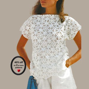 Op de afbeelding: Een wit shirt met korte mouwen gemaakt van ingewikkeld bloemvormig haakwerk met een boothals. Een ronde zwarte badge aan de linkerkant bevat de tekst 40% off on 2 or more patterns en een klein rood hartpictogram onder de gedrukte tekst.