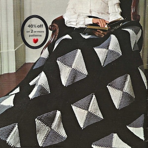 Pode incluir: Uma manta de malha preta com um padrão repetitivo de motivos em forma de diamante, cada um composto por dois segmentos triangulares contrastantes em cinza claro e cinza escuro. A manta está sobre uma cadeira, com um livro segurado por mãos.
