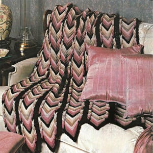 May include: A crocheted blanket with a chevron pattern in shades of black, pink, beige, and white is draped over a sofa. Two pink pillows rest on the sofa. The blanket's design creates a visual texture, adding warmth and style to the room.