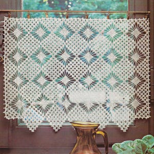 May include: Cream-colored, crocheted window curtain with a floral pattern. The curtain is rectangular and hangs from a brass rod. The design features repeating flower-like shapes and a scalloped edge. A glass pitcher sits below the curtain.