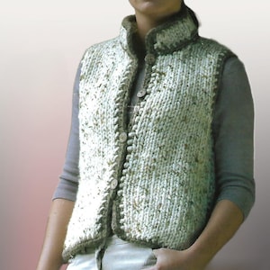 May include: A hand-knitted vest in cream and olive green. The vest features a high collar and a button-up front. The edges are trimmed with a darker olive green yarn. The chunky knit design adds texture and warmth to this cozy garment.