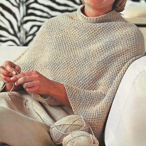 May include: A beige knitted poncho with a high collar. The poncho has a textured pattern and is being worked on with knitting needles and yarn. Two balls of yarn are visible in the foreground. The background features a zebra-striped pattern.