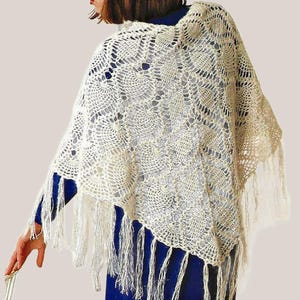 May include: A cream-coloured crochet shawl featuring an intricate pineapple-inspired openwork pattern. The garment is triangular in shape and finished with long, thin fringe tassels along the bottom edges, creating a textured and layered appearance.