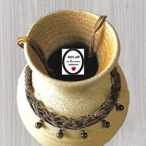 Puede incluir: Un collar de macramé marrón con intrincados patrones de cordón trenzado y cuentas esféricas de color marrón oscuro colgantes. El collar está colocado alrededor de un jarrón de cerámica beige texturizado. Una etiqueta blanca muestra 40% off on 2 or more patterns.