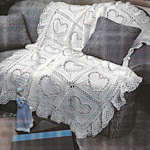 May include: A white, crocheted blanket with heart-shaped designs and a scalloped edge. The blanket is draped over a gray sofa, with two gray pillows. A small figurine in a blue dress is in the foreground.