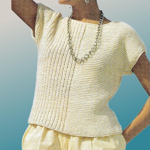 May include: Cream-colored, short-sleeved knit top with a boat neck. The top has a ribbed texture and a fitted silhouette. A silver chain necklace is draped around the neck. The model is wearing yellow pants.