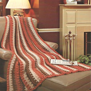 May include: A crocheted throw blanket featuring vertical stripes in shades of rust orange, dark brown, and cream. The blanket has a textured pattern with alternating stitch designs and is draped over a chair, with a newspaper resting on top.