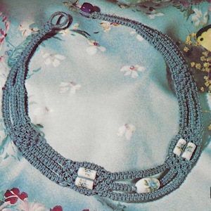 May include: A blue macrame necklace with decorative white and blue floral ceramic beads. The necklace has a woven design and a clasp closure. The necklace is displayed on a floral patterned background.