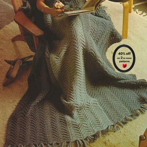 May include: A sage green knitted throw blanket with a chevron texture and fringe trim. The blanket features a circular graphic with the text 40% off on 2 or more patterns and a red heart icon. The blanket is draped over a wooden chair.