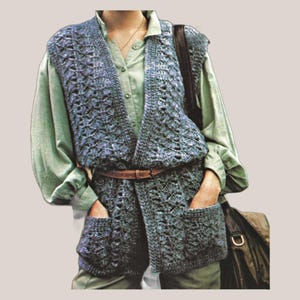 Pode incluir: Um colete azul de crochê com dois bolsos frontais e um cinto marrom. O colete é usado sobre uma camisa verde clara abotoada. Uma bolsa de couro marrom é jogada sobre o ombro.