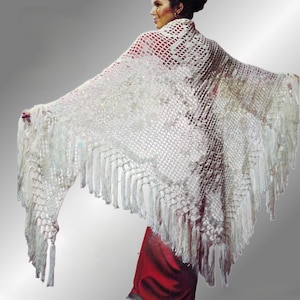 May include: A white, open-weave shawl with long fringe. The shawl is draped over the shoulders, with the arms extended. The garment is made of a delicate, possibly crocheted, material. The background is a gradient of gray.