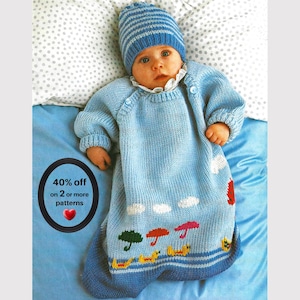 Könnte beinhalten: Ein hellblauer gestrickter Babyschlafsack mit passender gestreifter Mütze. Der Schlafsack hat einen weißen Spitzenkragen und eine untere Bordüre mit Intarsienmustern aus grünen, roten und rosa Regenschirmen, gelben Enten und weißen Wolken.