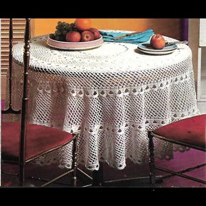 May include: A white, crocheted tablecloth with a detailed openwork pattern covers a round table. The tablecloth has a scalloped edge. A plate of fruit and an apple on a plate are on the table. Two red chairs are in the foreground.