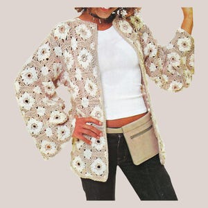 May include: A beige crochet jacket with white floral patterns. The jacket has long sleeves and a collarless design. The garment is worn over a white top and dark jeans. A beige fanny pack is worn around the waist.