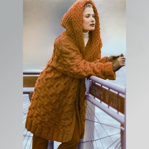 Può includere: Un lungo cappotto con cappuccio a maglia a trecce in una calda tonalità arancione terrosa. Il cappotto presenta un motivo strutturato e un ampio cappuccio. Le maniche sono lunghe e il cappotto sembra essere realizzato con un filato spesso e accogliente.