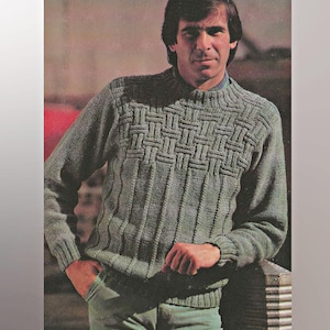 Könnte beinhalten: Ein gestrickter grüner Pullover mit einem strukturierten Korbgeflechtmuster auf der Brust und vertikalen Rippen am Körper. Der Pullover hat lange Ärmel und einen Rundhalsausschnitt. Die Person trägt eine grüne Hose.