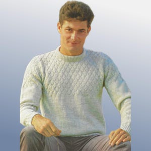 May include: A pale blue knitted sweater featuring a diamond pattern. The sweater has a crew neck and long sleeves with ribbed cuffs. The person is wearing grey trousers.