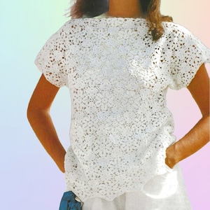May include: A white, short-sleeved crochet top with a floral pattern. The top has a round neckline and a loose fit. The crochet work is detailed and creates an open, airy design. The top is paired with white trousers.