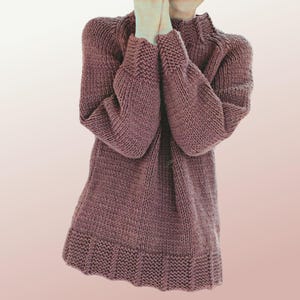 May include: A mauve-colored, hand-knitted sweater with a high neckline and long sleeves. The sweater has a ribbed hem and cuffs, and a slightly flared shape. The knit is a chunky, textured stitch, giving the garment a cozy appearance. The sleeves are wide and the overall style is relaxed.